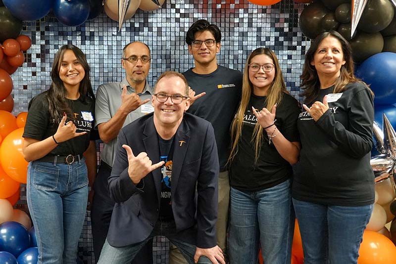 Six attendees pose together in front of a decorative backdrop with orange, blue, black, and silver balloons during a UTEP Engineering homecoming gathering, making hand gesture signs while wearing departmental apparel and event name badges.